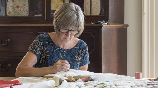 A woman with short grey hair and glasses is working on some embroidery at a table.